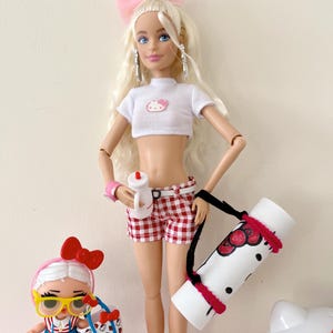 May include: A blonde doll with a pink bow in her hair is wearing a white crop top with a Hello Kitty design, red and white checkered shorts, and a black strap carrying a white yoga mat with a Hello Kitty design. A smaller doll with a pink bow and yellow glasses is holding a Hello Kitty bag.