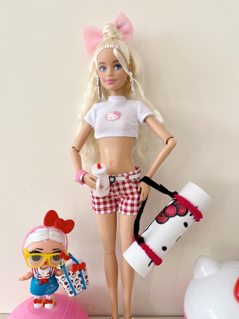 May include: A blonde doll with a pink bow in her hair is wearing a white crop top with a Hello Kitty design, red and white checkered shorts, and a black strap carrying a white yoga mat with a Hello Kitty design. A smaller doll with a pink bow and yellow glasses is holding a Hello Kitty bag.
