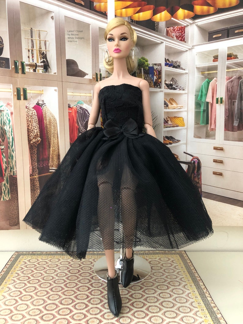 以下が含まれることがあります： 黒いストラップレスのドレスは、チュールスカートとウエストに黒いリボンが付いています。ドレスは、ブロンドの髪と青い目のファッションドールが着用しています。ドールは黒いヒールブーツを履いています。