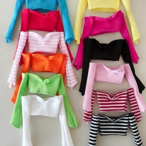 Peut inclure: Une collection de hauts épaules nues pour poupées de différentes couleurs et motifs. Les hauts comprennent des couleurs unies comme le bleu, le rouge, le jaune, le noir et le blanc, ainsi que des motifs rayés roses et blancs, et rouges et blancs. Les hauts ont des manches longues.