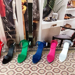 Op de afbeelding: Zeven paar sandalen met hoge hakken en doorzichtige hakken. De sandalen zijn in verschillende kleuren, waaronder rood, zwart, groen, blauw, roze, wit en nude.