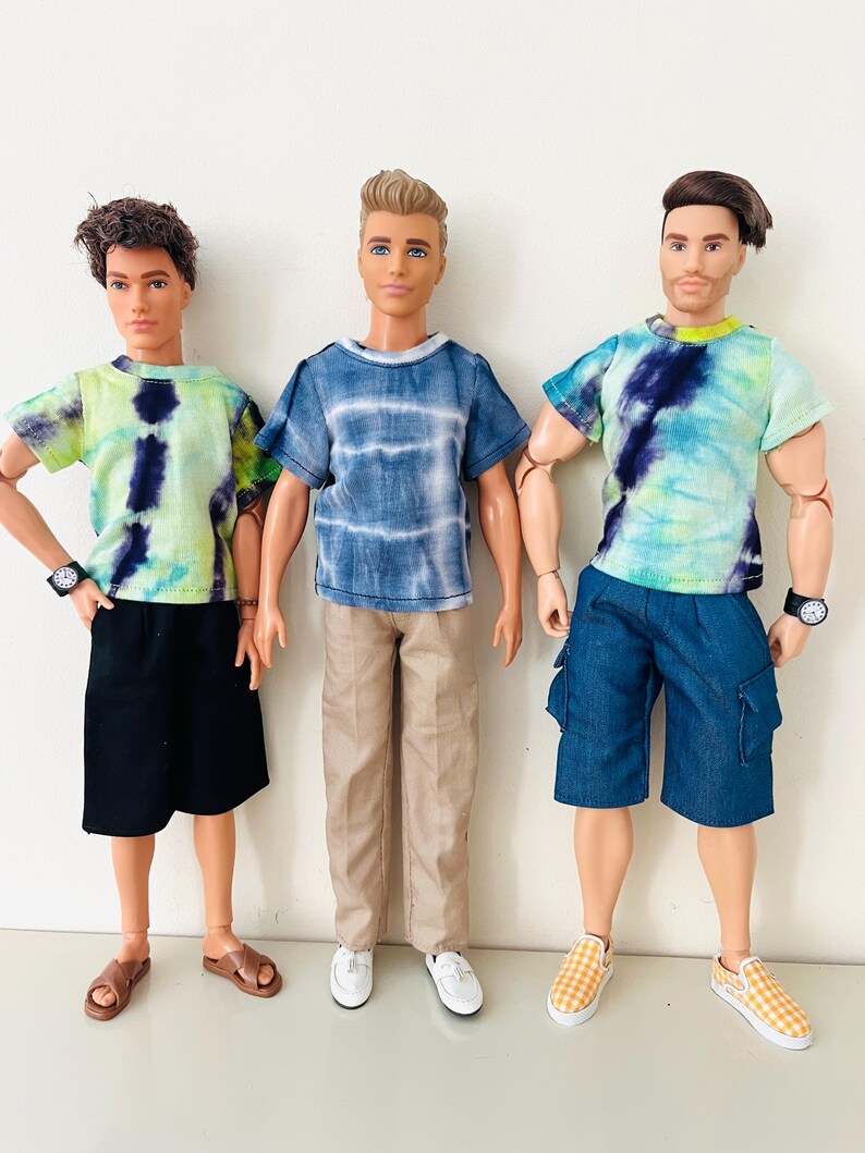 Puede incluir: Tres mu&ntilde;ecos masculinos de pie uno al lado del otro. El mu&ntilde;eco de la izquierda lleva una camiseta tie-dye verde lima y azul marino, pantalones cortos negros y sandalias marrones. El mu&ntilde;eco central lleva una camiseta tie-dye azul, pantalones beige y zapatos blancos. El mu&ntilde;eco de la derecha lleva una camiseta tie-dye verde lima y azul marino, pantalones cortos cargo azules y zapatos sin cordones a cuadros amarillos.