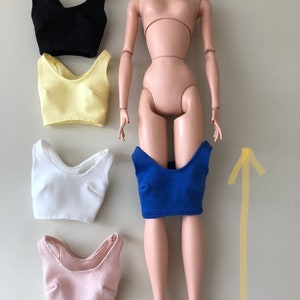 May include: A doll with light skin tone and blonde hair, accompanied by five sleeveless crop tops in black, pale yellow, white, light pink, and royal blue. The blue top is positioned on the doll's lower torso, while the others are arranged to the left.