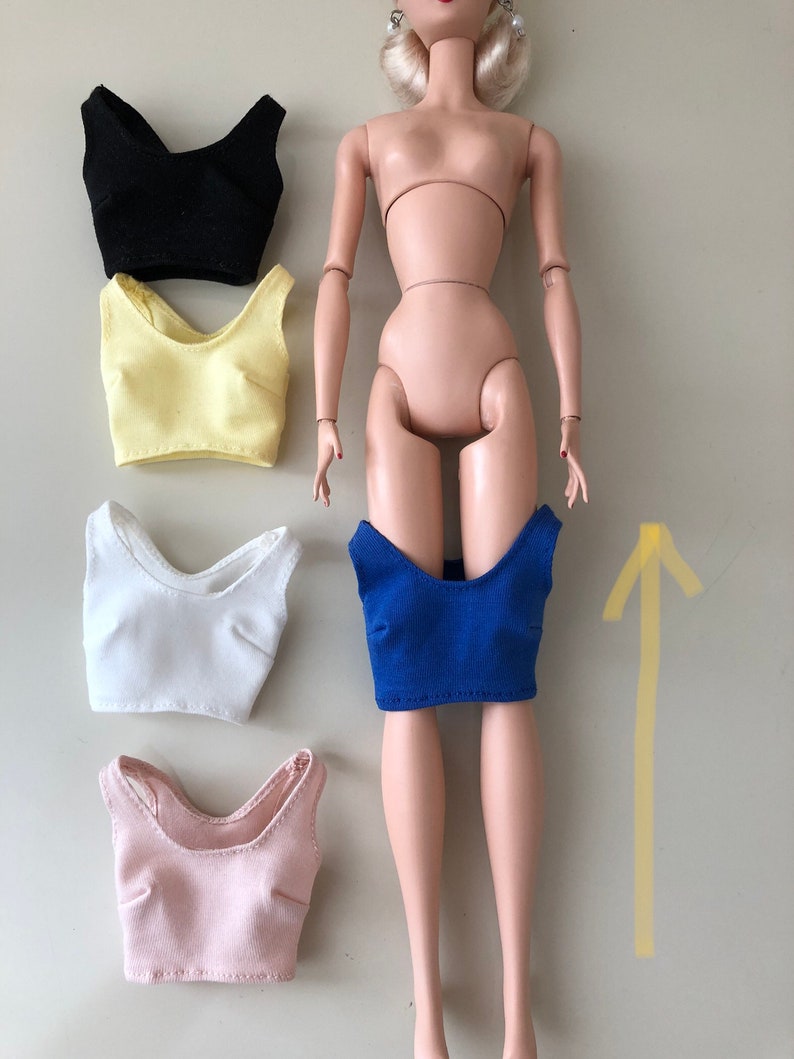 May include: A doll with light skin tone and blonde hair, accompanied by five sleeveless crop tops in black, pale yellow, white, light pink, and royal blue. The blue top is positioned on the doll's lower torso, while the others are arranged to the left.