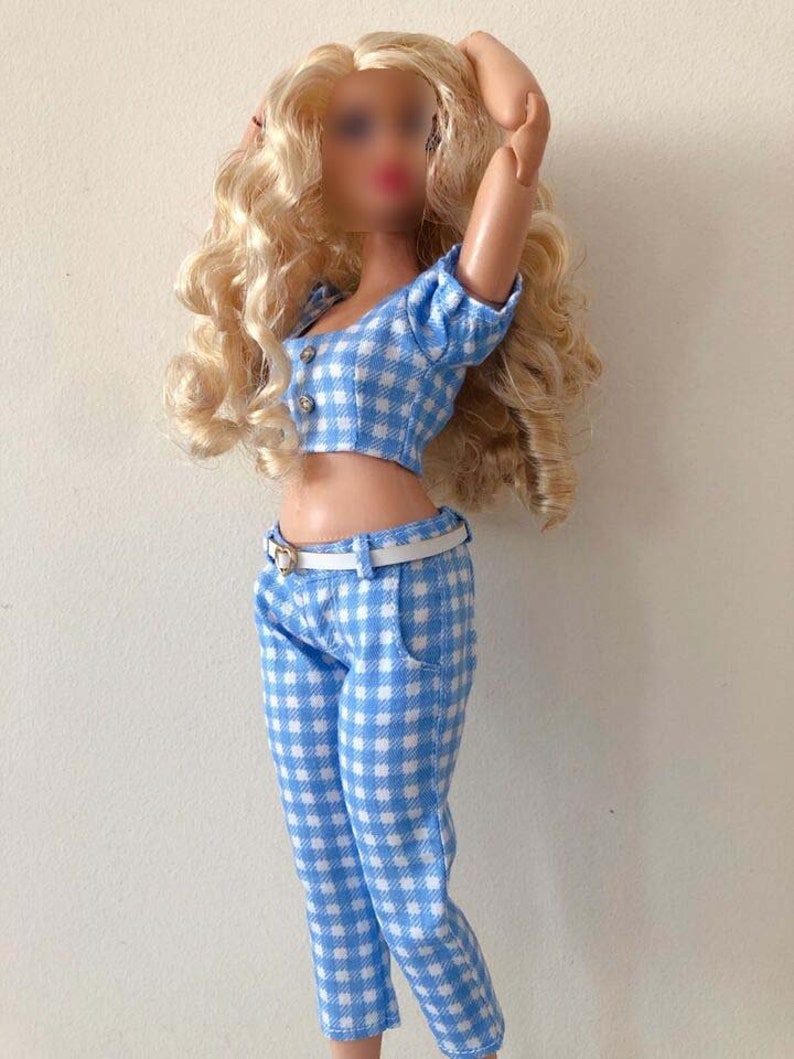 May include: A doll with long blonde curly hair wearing a two-piece outfit consisting of a light blue and white gingham crop top with two buttons and matching gingham pants. The outfit is accessorized with a thin white belt featuring a heart-shaped buckle.