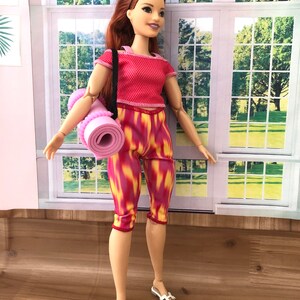May include: A doll wearing a red and white workout outfit with a pink yoga mat. The doll has long red hair and is wearing white sandals.
