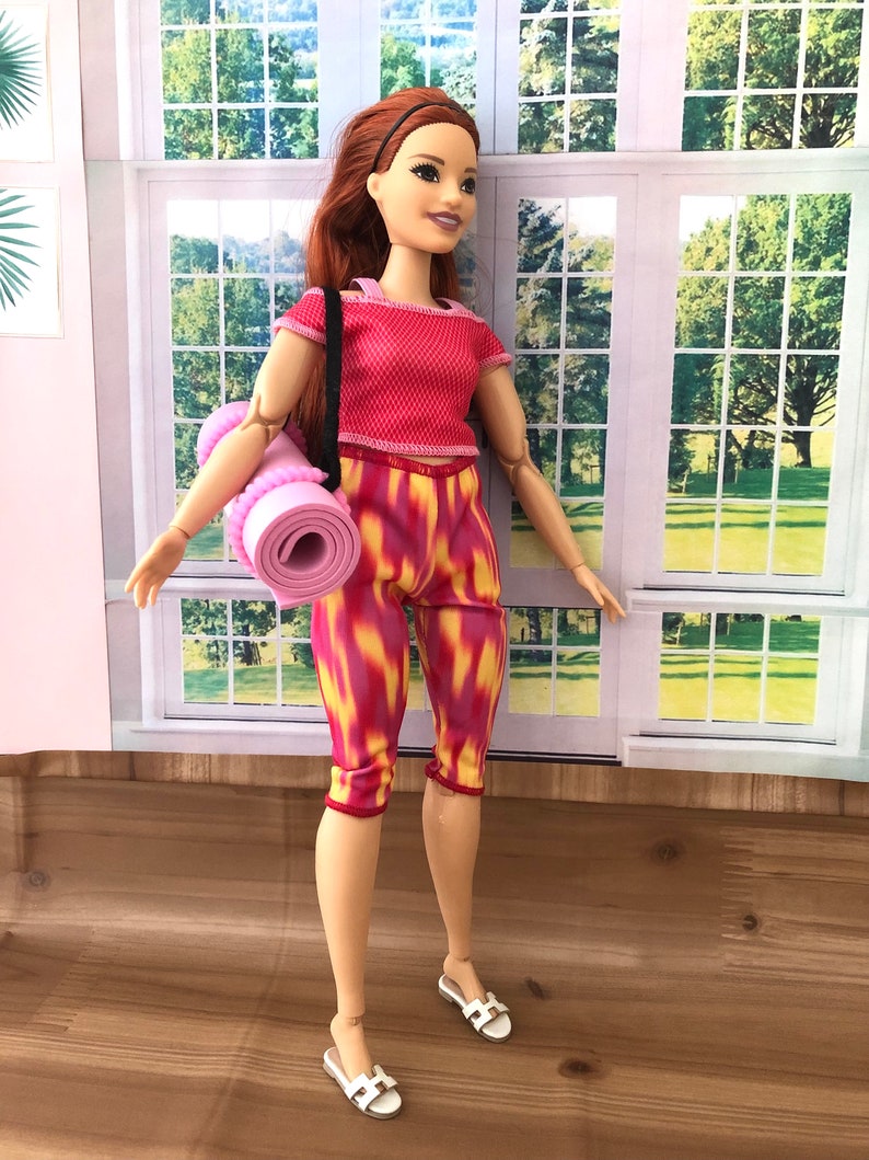 May include: A doll wearing a red and white workout outfit with a pink yoga mat. The doll has long red hair and is wearing white sandals.