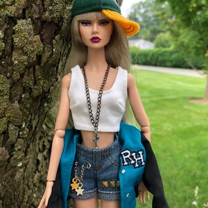 May include: A fashion doll with long blonde hair wearing a green and yellow baseball cap, a white crop top, denim shorts with gold star charms, and a teal and black varsity jacket with the letters RH embroidered on the chest and a cross necklace.