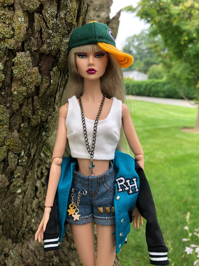 May include: A fashion doll with long blonde hair wearing a green and yellow baseball cap, a white crop top, denim shorts with gold star charms, and a teal and black varsity jacket with the letters RH embroidered on the chest and a cross necklace.