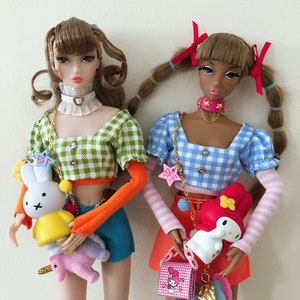 May include: Two fashion dolls with brown hair, one wearing a green gingham crop top with orange sleeves and blue shorts, the other in a blue gingham crop top with striped sleeves and a red skirt, both holding small character charms labeled BFF.