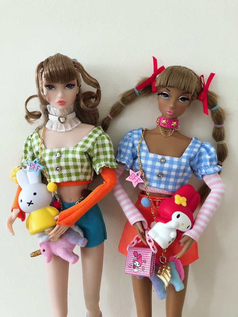 May include: Two fashion dolls with brown hair, one wearing a green gingham crop top with orange sleeves and blue shorts, the other in a blue gingham crop top with striped sleeves and a red skirt, both holding small character charms labeled BFF.