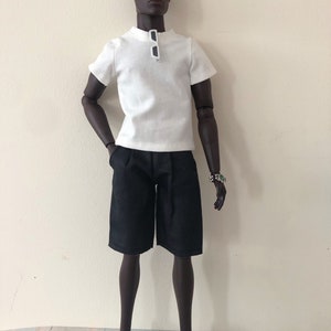 Puede incluir: Una mu&ntilde;eca articulada de piel oscura que viste una camiseta blanca de manga corta con cuello redondo, con gafas de sol blancas colgadas del cuello, pantalones cortos negros hasta la rodilla y mocasines dorados. Lleva un reloj plateado en la mu&ntilde;eca.