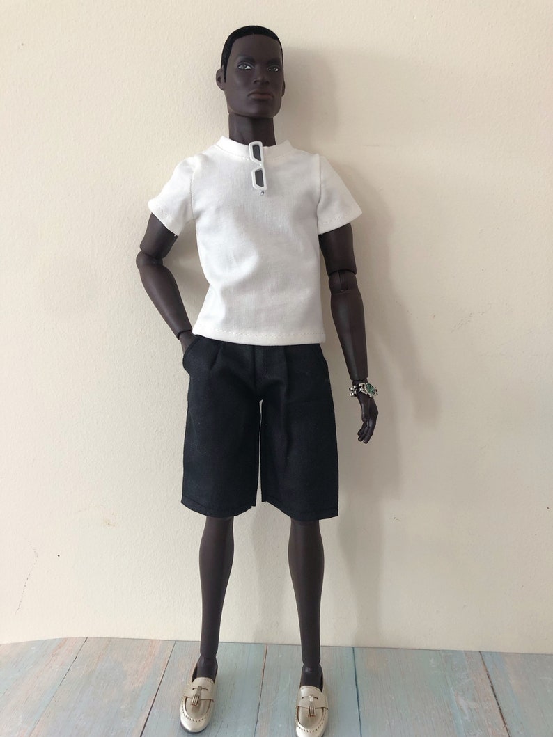 Puede incluir: Una mu&ntilde;eca articulada de piel oscura que viste una camiseta blanca de manga corta con cuello redondo, con gafas de sol blancas colgadas del cuello, pantalones cortos negros hasta la rodilla y mocasines dorados. Lleva un reloj plateado en la mu&ntilde;eca.