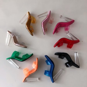 Könnte beinhalten: Acht Paar transparente Sandalen mit Absatz und unterschiedlichfarbigen Riemen. Die Riemen sind orange, grün, blau, schwarz, rot, rosa und zwei Brauntönen.