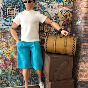 Puede incluir: Un mu&ntilde;eco con cabello casta&ntilde;o y gafas de sol negras que viste una camiseta blanca, pantalones cortos azul brillante y mocasines blancos. El mu&ntilde;eco est&aacute; junto a dos cajas de madera apiladas y un barril cil&iacute;ndrico de color canela.