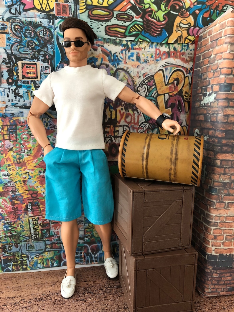 Puede incluir: Un mu&ntilde;eco con cabello casta&ntilde;o y gafas de sol negras que viste una camiseta blanca, pantalones cortos azul brillante y mocasines blancos. El mu&ntilde;eco est&aacute; junto a dos cajas de madera apiladas y un barril cil&iacute;ndrico de color canela.