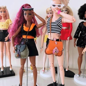 May include: A group of fashion dolls standing on display stands. One doll has long magenta hair, a black and orange crop top, a black skirt, and a green shoulder bag. Another doll has blonde hair, a black and white striped top, and an orange bag.