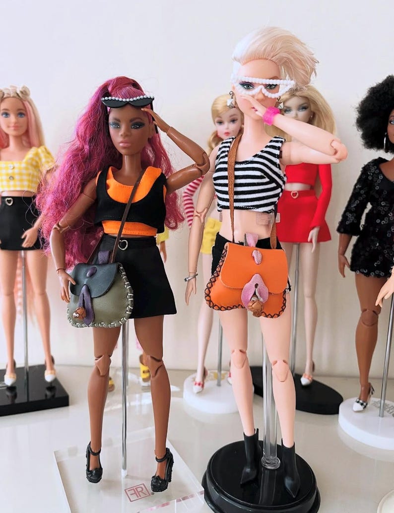 May include: A group of fashion dolls standing on display stands. One doll has long magenta hair, a black and orange crop top, a black skirt, and a green shoulder bag. Another doll has blonde hair, a black and white striped top, and an orange bag.
