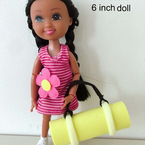 May include: A 6 inch doll with black hair in braids, wearing a pink and white striped dress with a pink flower on the front. The doll is holding a yellow yoga mat with black straps.