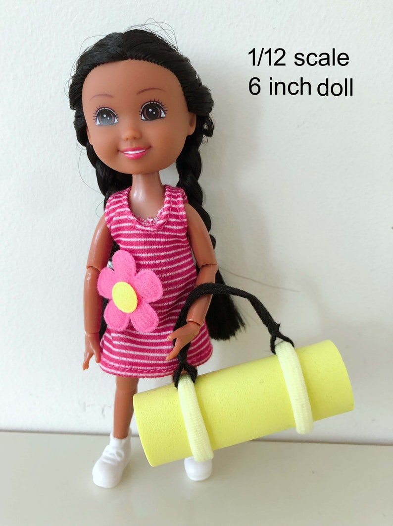 May include: A 6 inch doll with black hair in braids, wearing a pink and white striped dress with a pink flower on the front. The doll is holding a yellow yoga mat with black straps.