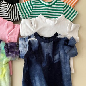 Puede incluir: Una colecci&oacute;n de camisetas peque&ntilde;as para mu&ntilde;ecas en varios colores y patrones, incluyendo una camiseta azul oscuro con efecto tie-dye, una camiseta blanca lisa, una a rayas verdes y blancas, una a rayas naranjas y blancas, una negra, una rosa y una tie-dye.
