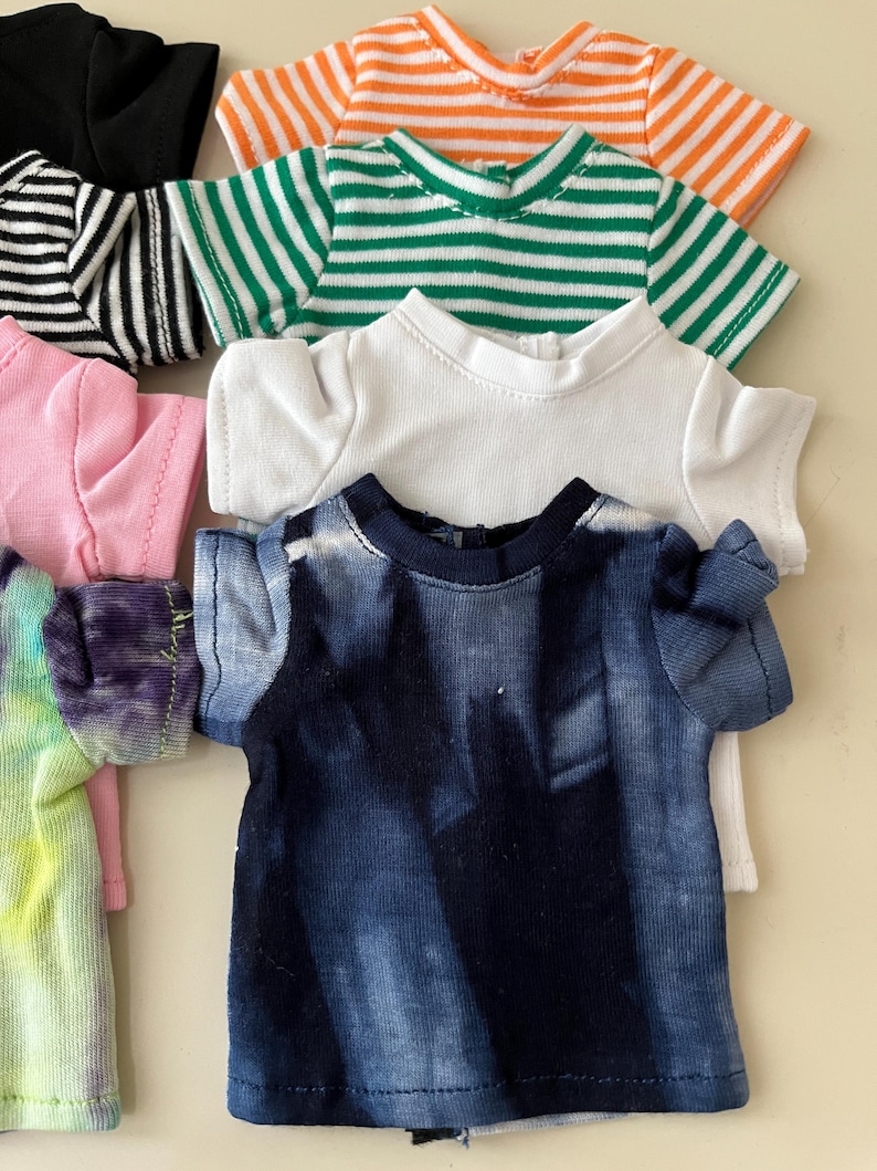 Puede incluir: Una colecci&oacute;n de camisetas peque&ntilde;as para mu&ntilde;ecas en varios colores y patrones, incluyendo una camiseta azul oscuro con efecto tie-dye, una camiseta blanca lisa, una a rayas verdes y blancas, una a rayas naranjas y blancas, una negra, una rosa y una tie-dye.