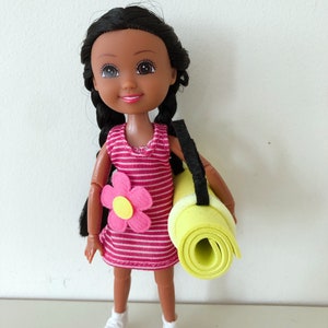 May include: A doll with dark brown hair in pigtails wearing a pink and white striped dress with a pink flower on the front. The doll is holding a rolled up yellow yoga mat with a black strap.