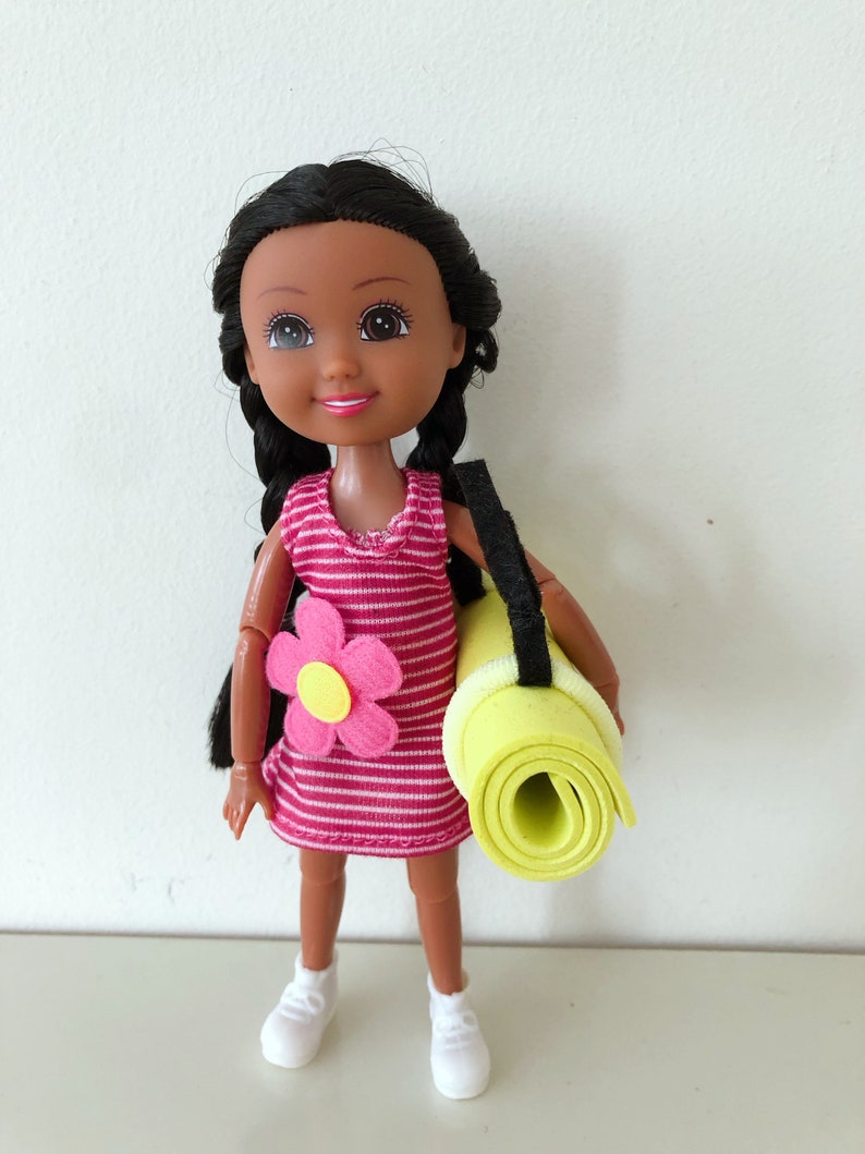 May include: A doll with dark brown hair in pigtails wearing a pink and white striped dress with a pink flower on the front. The doll is holding a rolled up yellow yoga mat with a black strap.