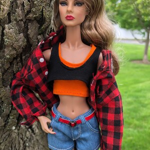 May include: A fashion doll with long wavy brown hair wearing a black and orange layered crop top, a red and black buffalo plaid button-down shirt, and blue denim shorts with a thin red belt featuring a small heart-shaped buckle at the waist.