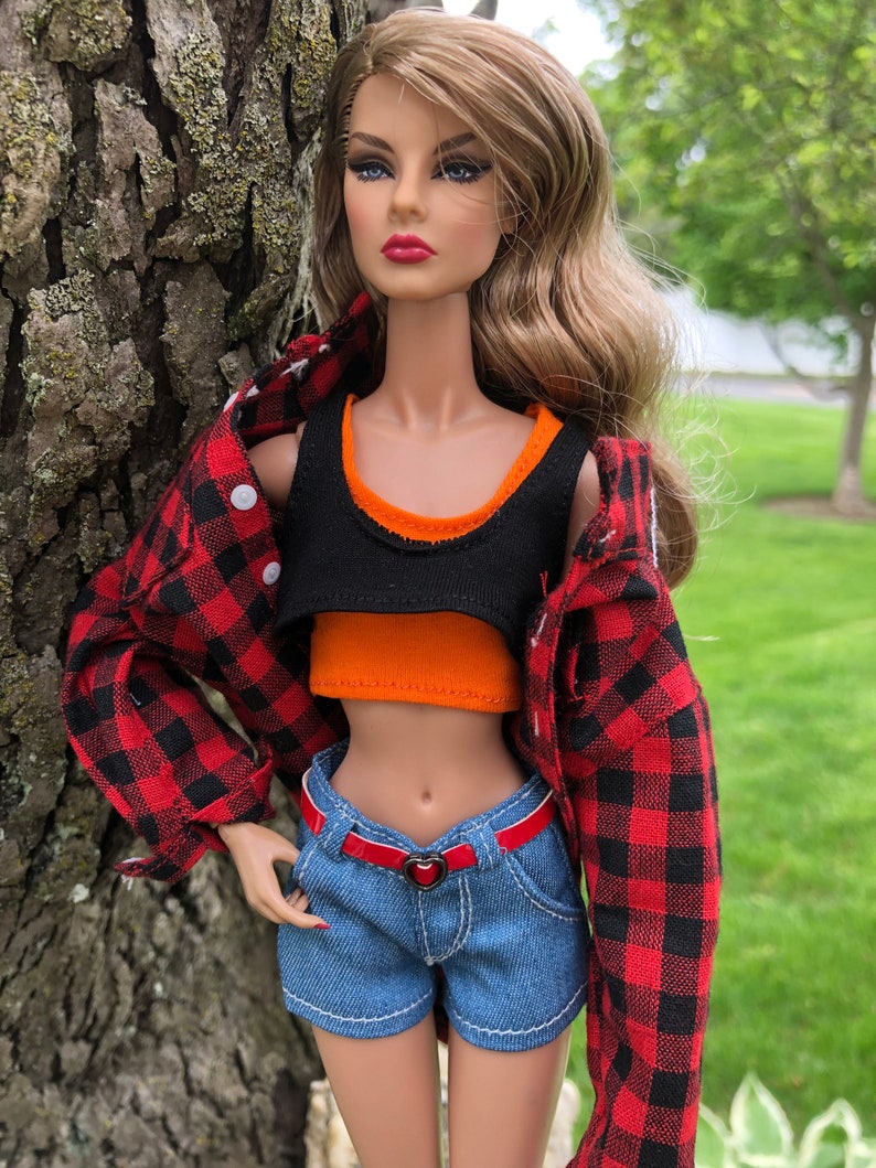 May include: A fashion doll with long wavy brown hair wearing a black and orange layered crop top, a red and black buffalo plaid button-down shirt, and blue denim shorts with a thin red belt featuring a small heart-shaped buckle at the waist.