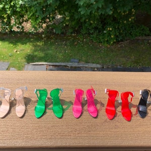 Op de afbeelding: Tien paar transparante sandalen met hak in verschillende kleuren, waaronder blauw, groen, roze, rood, zwart en wit. De sandalen zijn gerangschikt in twee rijen van vijf op een houten oppervlak.