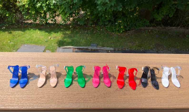 Op de afbeelding: Tien paar transparante sandalen met hak in verschillende kleuren, waaronder blauw, groen, roze, rood, zwart en wit. De sandalen zijn gerangschikt in twee rijen van vijf op een houten oppervlak.