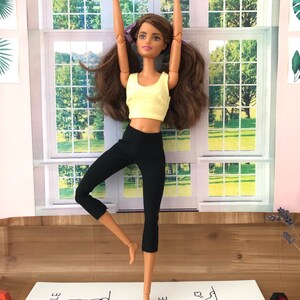 May include: A doll wearing a yellow tank top and black leggings is doing a yoga pose on a white yoga mat with the words "inhale" and "exhale" printed on it.