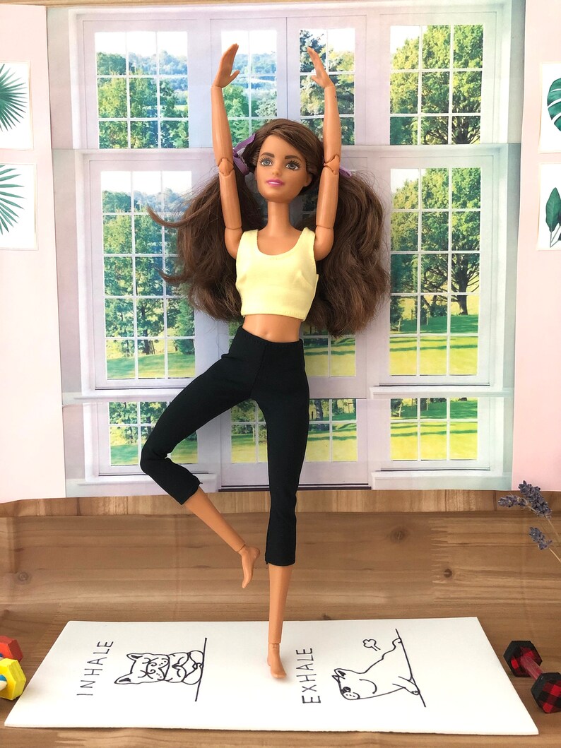 May include: A doll wearing a yellow tank top and black leggings is doing a yoga pose on a white yoga mat with the words "inhale" and "exhale" printed on it.