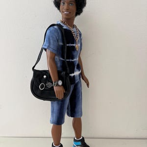 Puede incluir: Una mu&ntilde;eca de pelo oscuro rizado que viste una camiseta y pantalones cortos con estampado tie-dye en azul y negro. La mu&ntilde;eca lleva un collar de cadena dorada, un reloj de pulsera negro y un bolso de hombro negro con el logo plateado de Harley-Davidson. Zapatillas altas en negro, blanco y azul claro completan el conjunto.