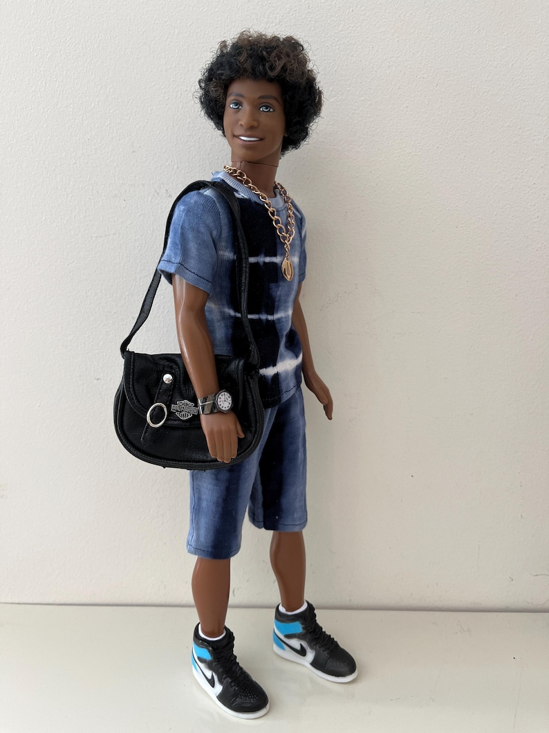 Puede incluir: Una mu&ntilde;eca de pelo oscuro rizado que viste una camiseta y pantalones cortos con estampado tie-dye en azul y negro. La mu&ntilde;eca lleva un collar de cadena dorada, un reloj de pulsera negro y un bolso de hombro negro con el logo plateado de Harley-Davidson. Zapatillas altas en negro, blanco y azul claro completan el conjunto.