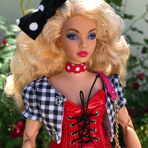 May include: A doll with blonde hair styled with a black and white polka dot bow. The doll wears a red vinyl corset-style top with black lacing, a red and white polka dot choker, and a black and white houndstooth cropped jacket with silver buttons.