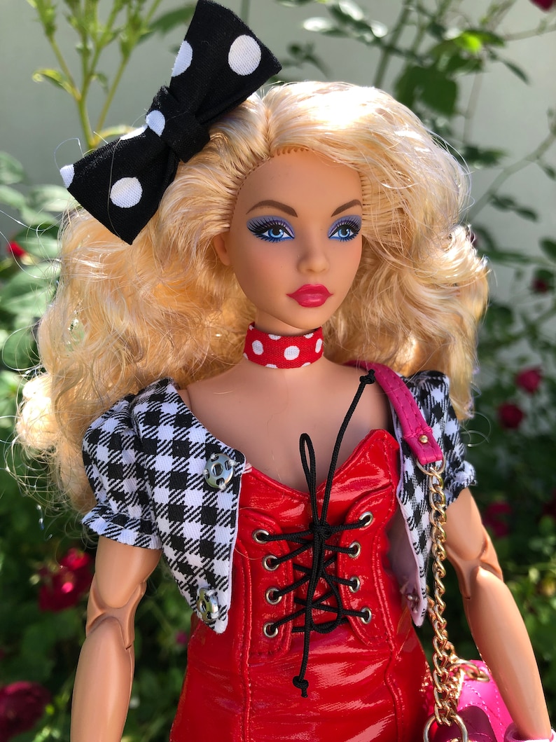 May include: A doll with blonde hair styled with a black and white polka dot bow. The doll wears a red vinyl corset-style top with black lacing, a red and white polka dot choker, and a black and white houndstooth cropped jacket with silver buttons.