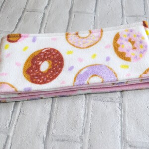 May include: A white fleece headband with a pink donut pattern. The donuts are decorated with sprinkles and have different coloured icings. The headband is folded in half.