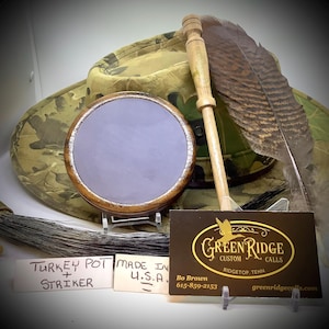 May include: A wooden turkey pot and striker with a gray slate surface. The pot is made in the USA. The image also includes a turkey feather, a wooden stick, and a business card for Green Ridge Custom Calls.