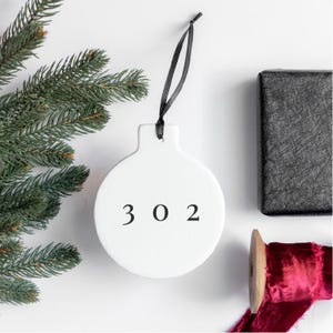 May include: White ceramic Christmas ornament with a black string for hanging. The ornament is round and has the number 302 printed on it in black.