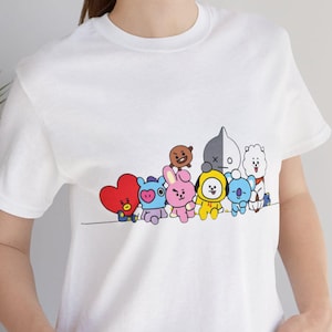 Könnte beinhalten: Weißes T-Shirt mit einem grafischen Druck einer Gruppe von Comicfiguren. Die Figuren sind alle in verschiedenen Farben und haben verschiedene Gesichtsausdrücke. Die Figuren tragen alle verschiedene Outfits. Das Shirt ist ein Rundhals-Shirt und hat kurze Ärmel.
