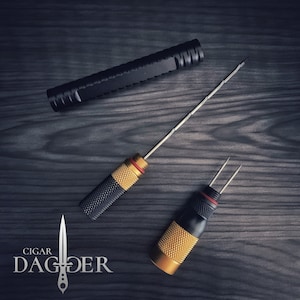 May include: Two cigar punch tools with gold and black accents. The tools are on a dark wood surface. The larger tool has a long, thin needle. The smaller tool has two prongs. The tools are labeled "CIGAR DAGGER".