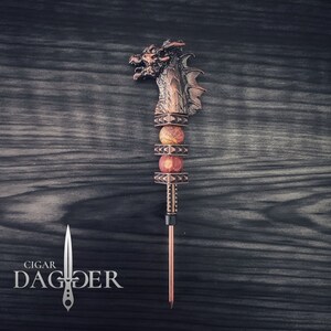 May include: A copper cigar dagger with a dragon head. The handle has red and tan stone beads. The text "CIGAR DAGGER" is in white. The dagger is designed for cigars.