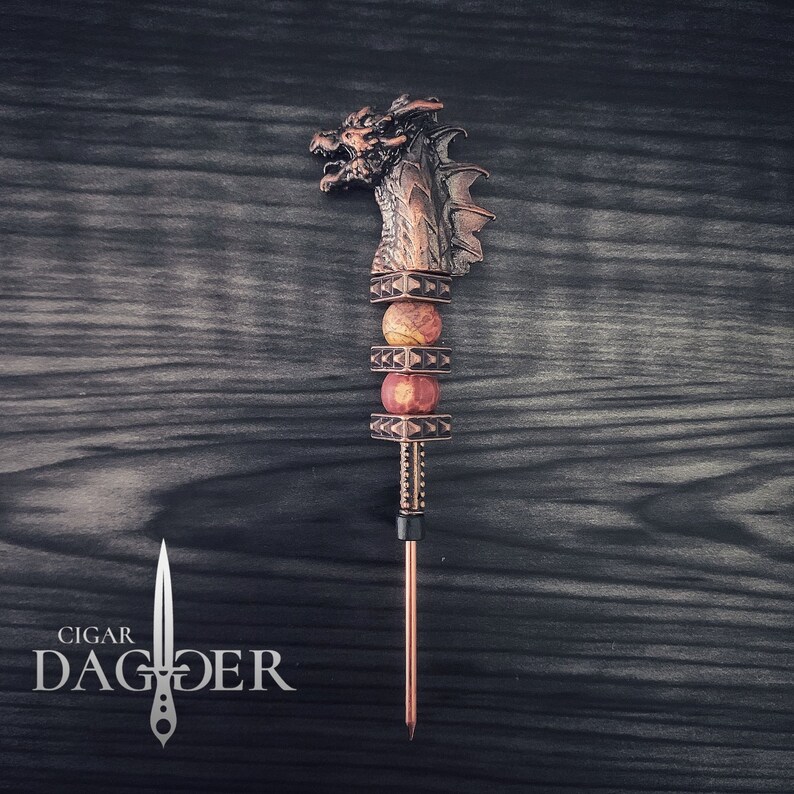 May include: A copper cigar dagger with a dragon head. The handle has red and tan stone beads. The text "CIGAR DAGGER" is in white. The dagger is designed for cigars.