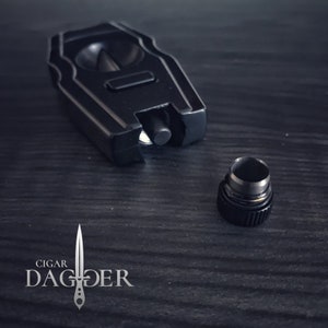 May include: A black cigar cutter with a silver trim and a textured design. The cutter is open and the blade is visible. The cutter is sitting on a dark wooden surface. The cutter has a small, round, black cap.