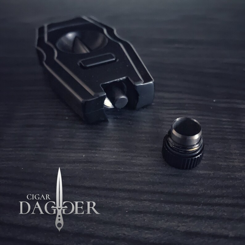 May include: A black cigar cutter with a silver trim and a textured design. The cutter is open and the blade is visible. The cutter is sitting on a dark wooden surface. The cutter has a small, round, black cap.