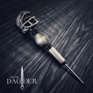 May include: A silver cigar poker with a football and football helmet design. The poker is made of metal and has a black cord wrapped around it. The helmet has a skull design. The poker is on a dark wood surface. The text "CIGAR DAGGER" is visible in the lower left corner of the image.