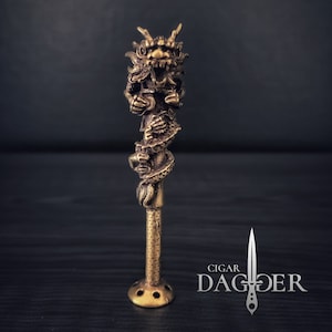May include: A gold-colored metal dragon cigar holder. The dragon is coiled around a long, thin tube with a round base. The dragon's head is at the top of the tube and has an open mouth.