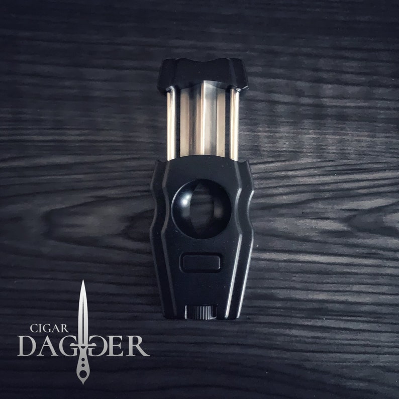 May include: A black cigar cutter with a silver blade and a silver knob. The cutter is shaped like a dagger and has the words "CIGAR DAGGER" printed on the bottom.